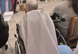 Dzieci w strojach jasełkowych występują przed seniorami siedzącymi na krzesłach i wózku