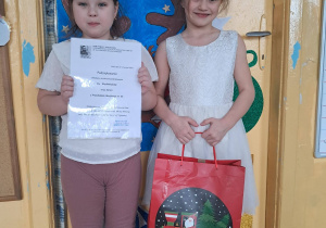 Dwie dziewczynki stoją przy drzwiach, jedna trzyma dyplom, druga świąteczną torbę prezentową.