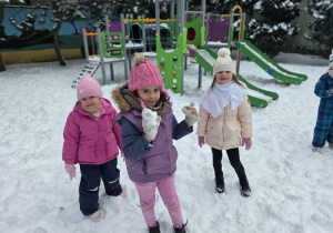 Dzieci w zimowych ubraniach stoją na zaśnieżonym placu zabaw i trzymają śnieg w rękach.