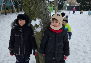 Dwoje dzieci w kombinezonach stoi przy drzewie na ośnieżonym podwórku.