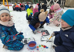 Grupa dzieci siedzi na śniegu i wspólnie maluje kolorowe wzory.