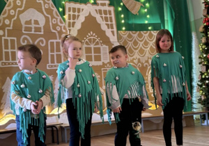 Czworo dzieci w zielonych strojach stoi na scenie przed świąteczną dekoracją.