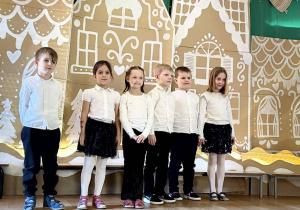 Dzieci w białych koszulach stoją przed dekoracją świąteczną.