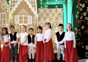 Dzieci w strojach stoją na scenie obok choinki.