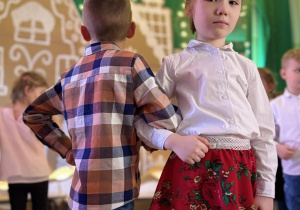 Dwoje dzieci stoi na scenie, jedno odwrócone tyłem, drugie patrzy w stronę aparatu.