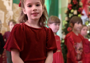 Dziewczynka w czerwonej sukience stoi na scenie, w tle inne dzieci.