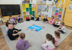 Dzieci siedzą w kole na dywanie i biorą udział w zajęciach dydaktycznych.