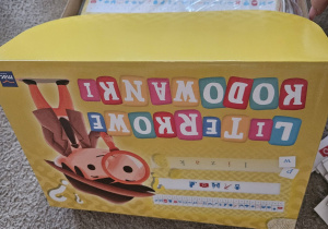 Pudełko z grą edukacyjną „Literkowe kodowanki” leży na dywanie w sali.
