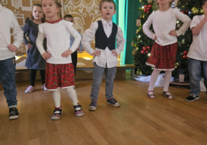 Grupa dzieci tańczy na scenie przy choince podczas występu świątecznego.