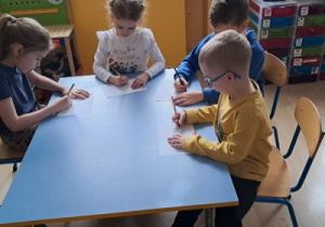 Dzieci siedzą przy stoliku i rysują na kartkach papieru.