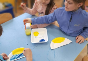 Dzieci malują farbami papierowe elementy przy stoliku.