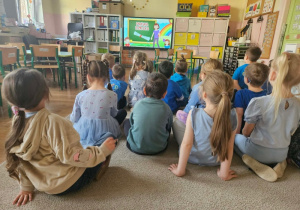 Dzieci siedzą na dywanie i oglądają prezentację o prawach dziecka na ekranie.