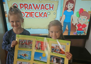 Dwie dziewczynki trzymają planszę „Prawa dziecka” na tle ekranu multimedialnego.