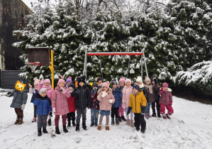 Grupa dzieci stoi na śniegu na placu zabaw i macha do zdjęcia.