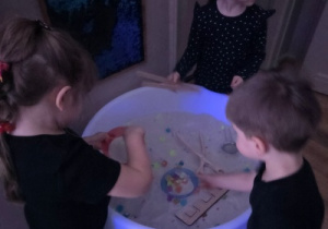 Troje dzieci bawi się przy podświetlanym stoliku z piaskiem sensorycznym i różnymi przedmiotami.