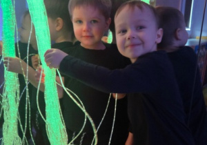 Dwoje dzieci stoi przy świecących światłowodach, obserwując kolory.