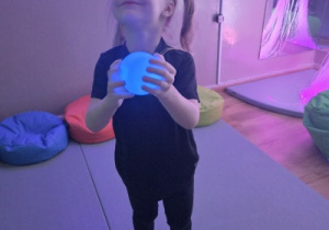 Dziewczynka trzyma świecącą piłkę i patrzy w górę, stojąc na miękkim materacu.