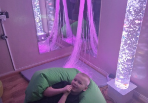 Dziecko odpoczywa na dużej pufie w sali sensorycznej, w otoczeniu delikatnego światła.