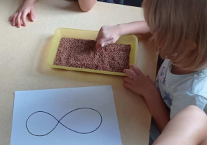 Dziewczynka palcem rysuje cyfrę osiem na tacy z kaszą.