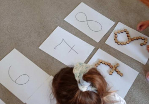 Dziewczynka układa na kartce, na dywanie obraz cyfry z drewnianych kółek.