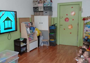 Dzieci siedzą na dywanie i oglądają na tablicy interaktywnej film edukacyjny o dokarmianiu ptaków zimą.