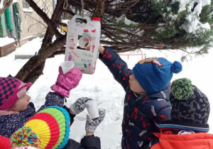 Dzieci przy pomocy plastikowych kubeczków nasypują ziarna do plastikowego karmnika.