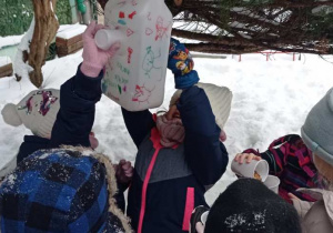 Dzieci stoją w ogrodzie przedszkolnym , otaczając drzewo z zawieszonym karmnikiem i wsypują do niego ziarna.