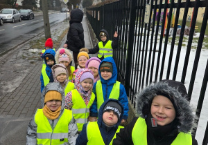Grupa przedszkolaków w kamizelkach odblaskowych idzie chodnikiem w stronę apteki pod opieką nauczyciela.