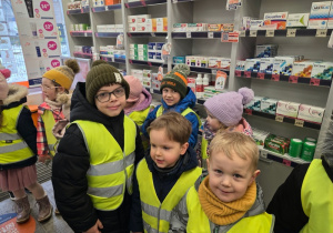 Dzieci stoją w aptece przy półkach z lekami i słuchają opowieści podczas zwiedzania.