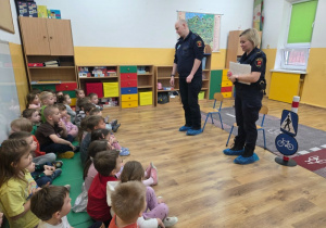 Funkcjonariusze Straży Miejskiej stoją przed grupą dzieci siedzących na dywanie w sali przedszkolnej. Trwa rozmowa o zasadach bezpieczeństwa.