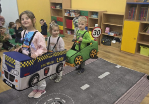 Dzieci uczestniczą w zabawie edukacyjnej – mają na sobie makiety pojazdów (radiowóz, samochód osobowy). Na podłodze rozłożona jest mata imitująca jezdnię z przejściem dla pieszych i znakami drogowymi.