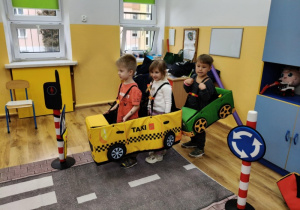 Troje dzieci w makietach samochodów stoi na macie przedstawiającej drogę. Obok ustawione są znaki drogowe i sygnalizator.