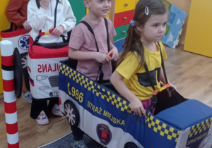 Przedszkolaki w makietach pojazdów (karetka i radiowóz Straży Miejskiej) biorą udział w zabawie edukacyjnej na macie przedstawiającej drogę.