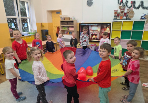Dzieci stoją w kole. Trzymają kolorową chustę. Na środku leżą czerwone balony w kształcie serc.