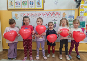 Dziewczynki stoją w rzędzie. Trzymają czerwone balony w kształcie serc.