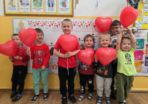 Chłopcy stoją obok siebie. Trzymają czerwone balony w kształcie serc i uśmiechają się.