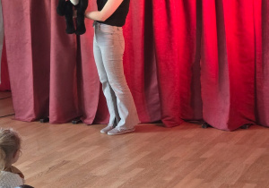 Aktorka trzyma czarną kukiełkę kota na tle czerwonej kurtyny, przed nią siedzą dzieci oglądające przedstawienie.