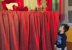 Chłopiec stoi przy scenie z czerwonymi zasłonami i patrzy na kukiełkę gąski wystającą zza dekoracji.