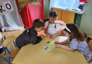 Troje dzieci siedzi przy stoliku i dopasowuje obrazki do mapy Polski.