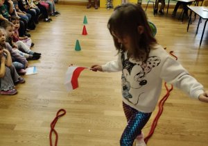 Dziewczynka przechodzi po linie trzymając małą flagę Polski. Inne dzieci siedzą i obserwują tor przeszkód.