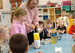 Nauczycielka wraz z grupą małych dzieci przygotowuje ciasto przy stole w sali przedszkolnej. Na stole znajdują się składniki, m.in. jajka, mąka i olej.