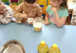 Dwoje dzieci miesza składniki w dzbanku podczas wspólnego przygotowywania ciasta przy stole.