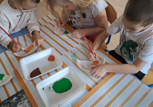 Dzieci malują farbami papierowe talerzyki podczas zajęć plastycznych o dinozaurach.