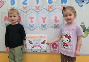 Dwoje dzieci stoi przed tablicą z napisem „Dzień Motyla”. Trzymają kartkę informującą o święcie motyli w przedszkolu.