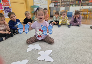 Dziewczynka siedzi na dywanie i pokazuje dwie niebieskie papierowe sylwety motyli. W tle widać grupę dzieci siedzących w kręgu.