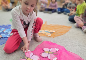 Dziewczynka układa różowe papierowe motyle na kolorowej chuście. W tle inne dzieci siedzą i obserwują zadanie.