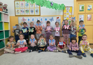 Grupa przedszkolaków siedzi i stoi w sali. Każde dziecko trzyma w rękach kolorowego papierowego motyla.