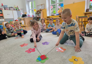 Dzieci siedzą na dywanie i uderzają łapkami w wskazane liczby.