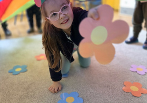 Dziewczynka w okularach czołga się pod kolorową chustą animacyjną i trzyma w ręku papierowy kwiatek. Na podłodze wokół niej leżą inne kolorowe kwiaty.