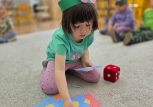 Dziewczynka siedzi na dywanie.Obok niej leży duża kostka do gry, której używa do zabawy matematycznej.